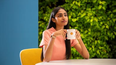 A person is holding a white coffee mug with the Shell logo, seated at a round table. The background features lush greenery, creating a refreshing and relaxed atmosphere.​