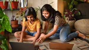 Two girls with dark hair sitting on a rug in a cozy indoor setting, focused on a laptop. A girl, wearing a yellow shirt with white patterns, uses the laptop, while another girl in a leopard print top smiles and leans in closer, pointing at the screen. Potted plants surround them, creating a cozy atmosphere. A second laptop is nearby.