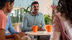 A close-up of a group conversation around a small table with bright orange coffee mugs. The setting includes greenery in the background, creating a relaxed and informal atmosphere. The person in the light green shirt appears to be speaking while others listen attentively.​