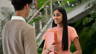 Two individuals are standing and conversing in a modern indoor space with greenery and a staircase in the background. One person is holding an orange laptop sleeve, suggesting a professional discussion or meeting.