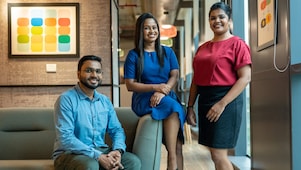 Three professionals with dark hair smiling and posing for a photo in an office. A man in a light blue shirt and glasses sits on a sofa, with two women standing beside him—one in a blue dress and the other in a red top and black skirt.