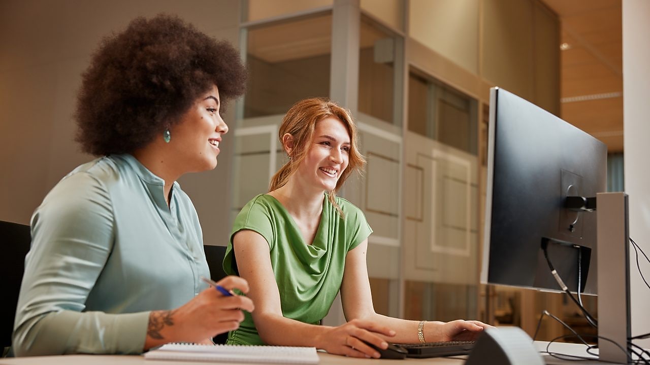 Two women are sitting at a desk in an office environment, one with dark curly hair wearing a light blue top and the other with straight blonde hair in a green top. They are smiling and engaged in a discussion while looking at a computer screen. A notebook and pen are placed on the table in front of them.