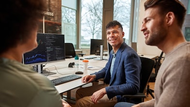 Three diverse people conversing in an office, with a computer monitor displaying graphs and codes in front of them. A man with dark curly hair in a blue blazer smiles at an out-of-focus woman with dark hair, while another man looks at her and appears to be engaged.