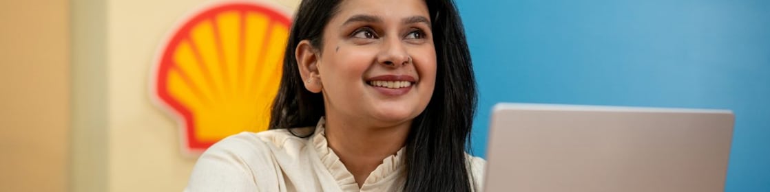 A person is working on a laptop at a desk with a Shell logo visible on the wall behind. A yellow notebook and pen are placed on the desk, suggesting a focused work environment. The background features vibrant blue and yellow panels.
