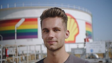 Person standing in front of a Shell storage tank displaying a rainbow banner and Shell logo