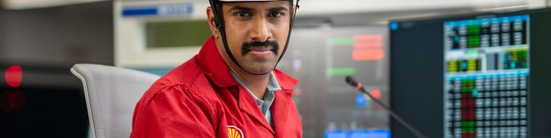 This image shows an individual wearing a white safety helmet and a red Shell-branded PPE, seated at a control desk. The person is positioned near large monitors displaying complex data and system metrics. The environment is well-lit and organized, emphasizing safety and operational efficiency.