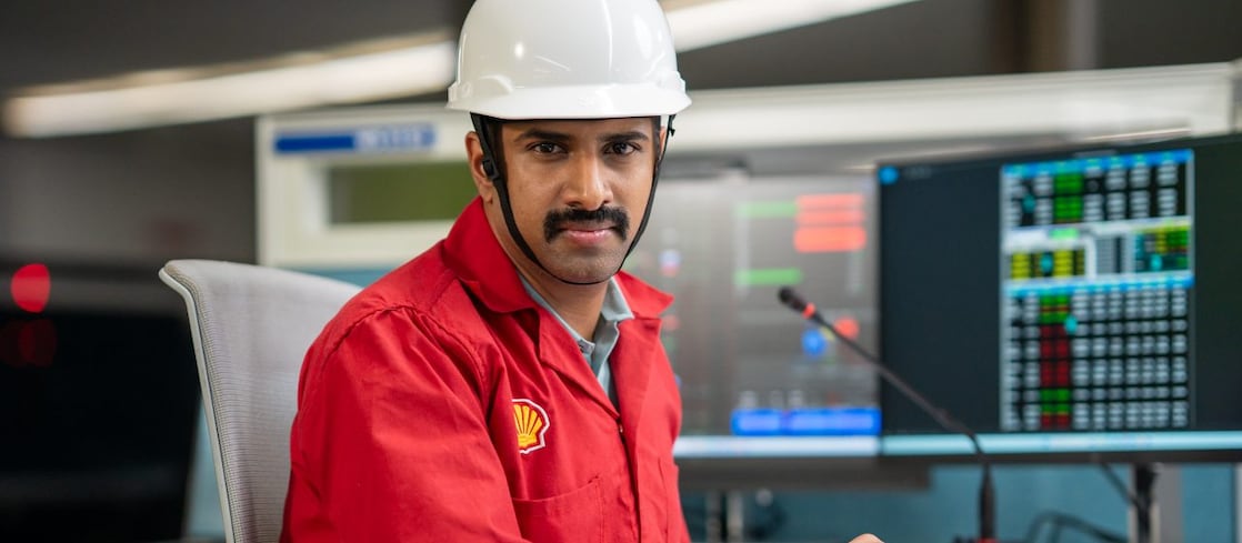 This image shows an individual wearing a white safety helmet and a red Shell-branded PPE, seated at a control desk. The person is positioned near large monitors displaying complex data and system metrics. The environment is well-lit and organized, emphasizing safety and operational efficiency.