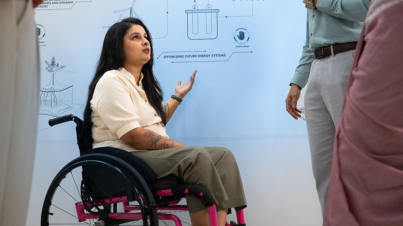 A closer view of the same discussion near the whiteboard. The person in the wheelchair is gesturing toward the diagrams, emphasizing a point about energy systems. The setting highlights collaboration and inclusivity in a technical environment.