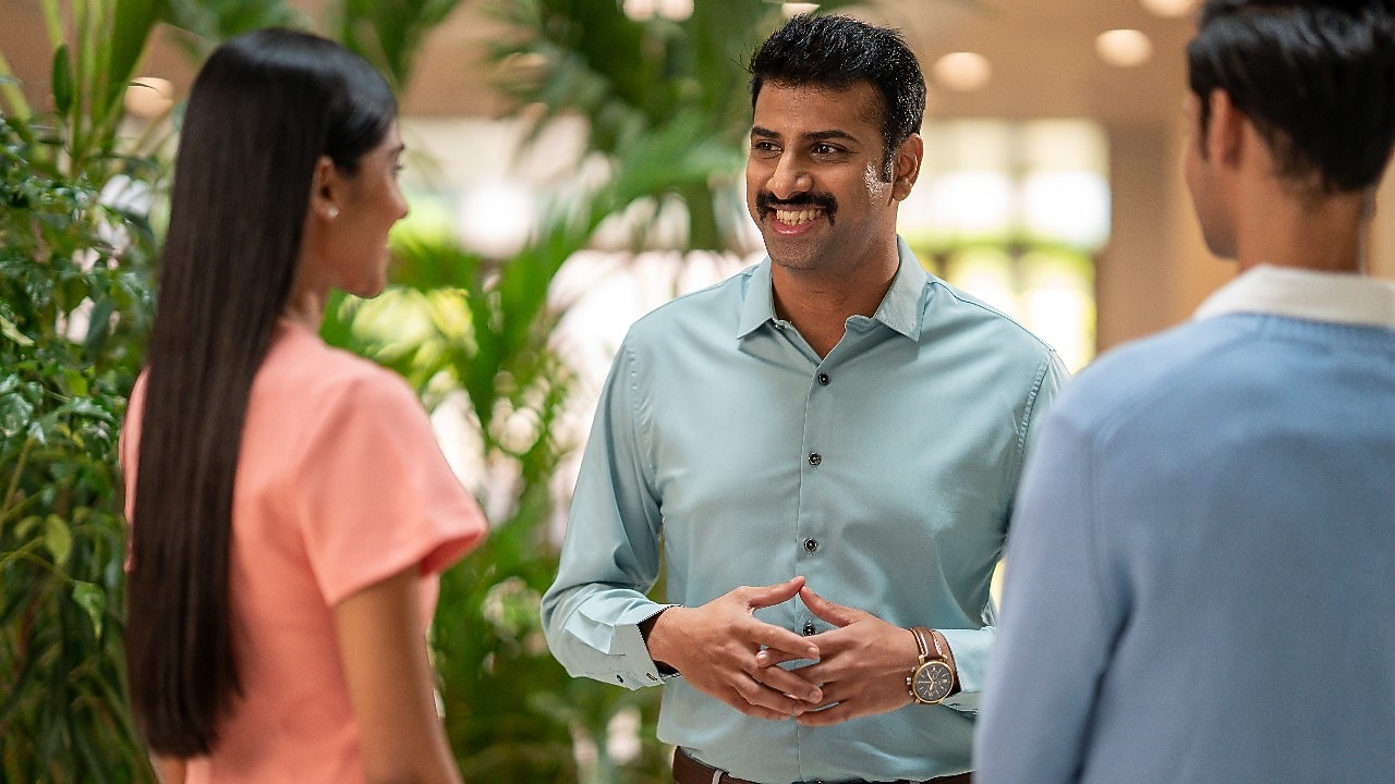 A person in a light green button-down shirt is engaged in conversation with two others. The setting appears to be an indoor space with lush green plants in the background, giving a natural and welcoming atmosphere. The individual in the center is using hand gestures, suggesting an active discussion or explanation.