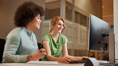 Two women are sitting at a desk in an office environment, one with dark curly hair wearing a light blue top and the other with straight blonde hair in a green top. They are smiling and engaged in a discussion while looking at a computer screen. A notebook and pen are placed on the table in front of them.