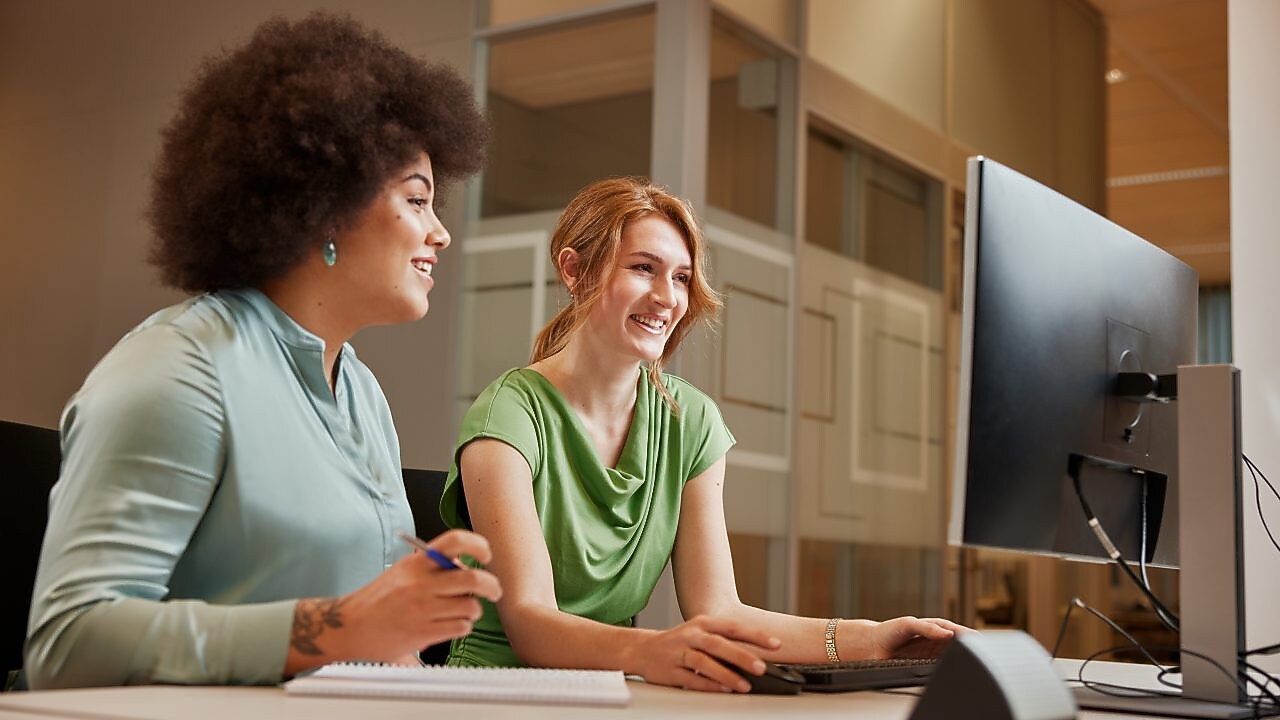 Two women are sitting at a desk in an office environment, one with dark curly hair wearing a light blue top and the other with straight blonde hair in a green top. They are smiling and engaged in a discussion while looking at a computer screen. A notebook and pen are placed on the table in front of them.