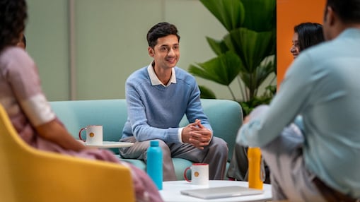 A person in a light blue sweater is seated in a lounge area, listening attentively to others. The setting includes colorful chairs, coffee mugs, and a relaxed atmosphere suitable for brainstorming or informal meetings.