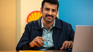A person is seated at a desk working on a laptop, with a Shell logo visible on the wall behind. The individual is holding a pen, indicating active engagement in work or note-taking.