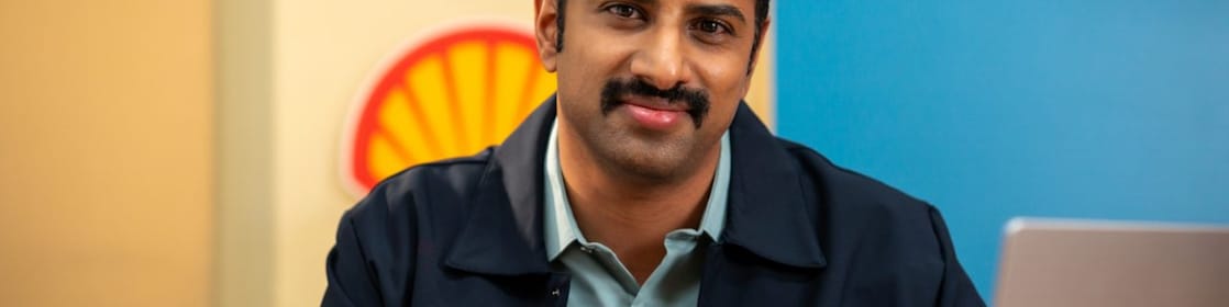 A person is seated at a desk working on a laptop, with a Shell logo visible on the wall behind. The individual is holding a pen, indicating active engagement in work or note-taking.