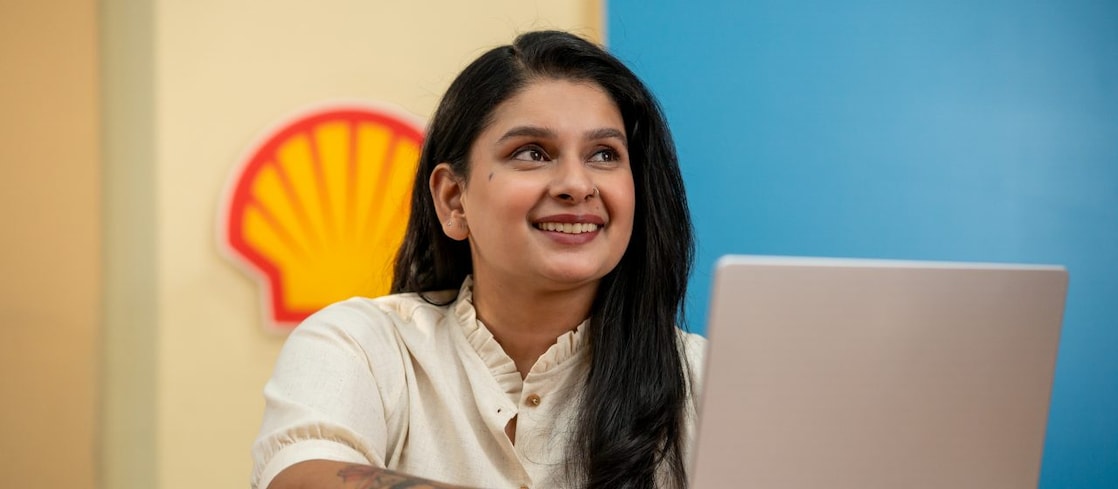A person is working on a laptop at a desk with a Shell logo visible on the wall behind. A yellow notebook and pen are placed on the desk, suggesting a focused work environment. The background features vibrant blue and yellow panels.