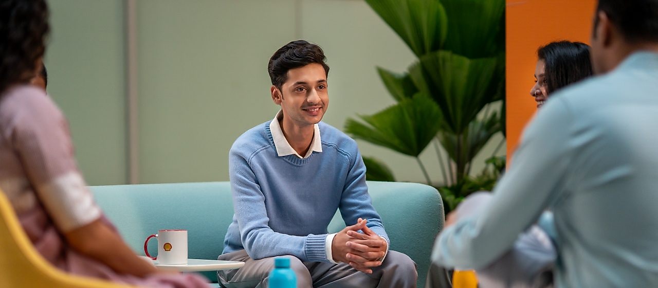 A person in a light blue sweater is seated in a lounge area, listening attentively to others. The setting includes colorful chairs, coffee mugs, and a relaxed atmosphere suitable for brainstorming or informal meetings.​