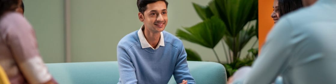 A person in a light blue sweater is seated in a lounge area, listening attentively to others. The setting includes colorful chairs, coffee mugs, and a relaxed atmosphere suitable for brainstorming or informal meetings.​