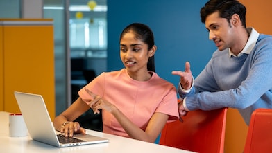Two individuals are seated at a desk with a laptop open, appearing to discuss something on the screen. The setting is colorful and modern, with orange and yellow panels in the background. A coffee mug is placed nearby, adding to the casual work atmosphere.​