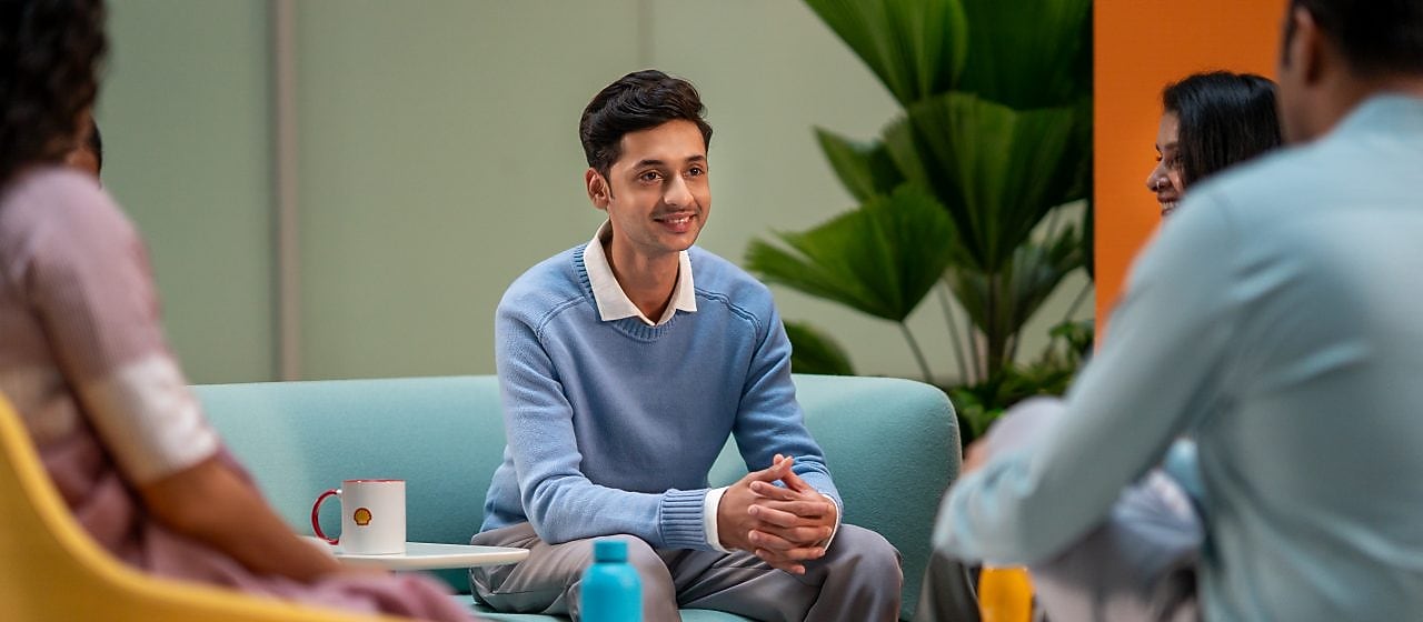 A person in a light blue sweater is seated in a lounge area, listening attentively to others. The setting includes colorful chairs, coffee mugs, and a relaxed atmosphere suitable for brainstorming or informal meetings.​