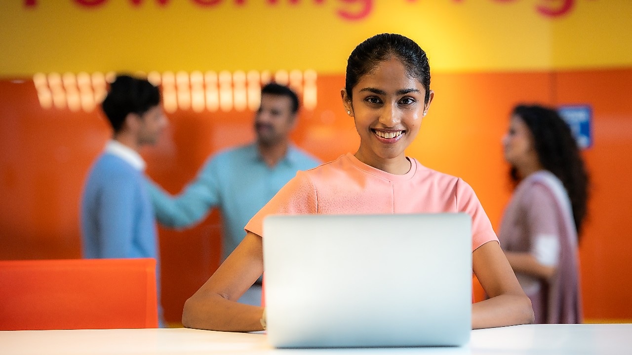 An individual is seated at a desk working on a laptop. The background shows the same orange wall with “Powering Progress” text and a group of people conversing, reinforcing the collaborative and energetic environment.​