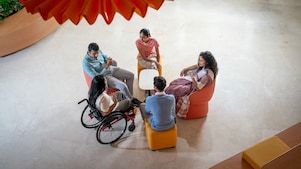 An overhead view of a small group discussion in a modern lounge area. The group is seated around a white table and chairs, including one person in a wheelchair. The space is open and well-lit, with a contemporary design and a large decorative element hanging above.​