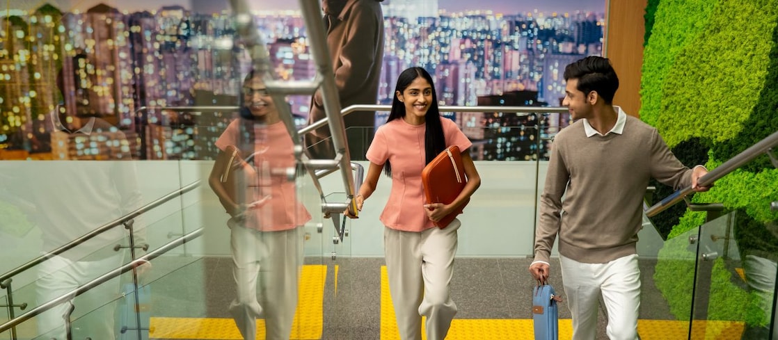 Two people walking down a modern indoor staircase with glass railings. One person is holding a folder, and the other is carrying a water bottle. The background features a large cityscape mural and a green wall decoration.” ​