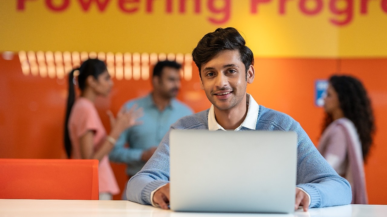 A person is working on a laptop in a vibrant workspace with an orange wall behind. In the background, others are engaged in conversation, creating a dynamic and interactive office environment.