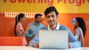 A person is working on a laptop in a vibrant workspace with an orange wall behind. In the background, others are engaged in conversation, creating a dynamic and interactive office environment.​