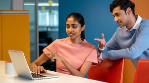 Two individuals are seated at a desk with a laptop open, appearing to discuss something on the screen. The setting is colorful and modern, with orange and yellow panels in the background. A coffee mug is placed nearby, adding to the casual work atmosphere.​