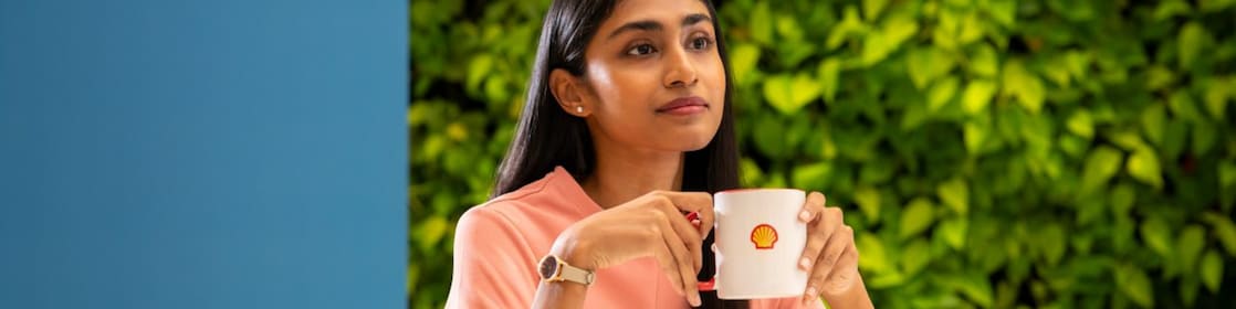 A person is holding a white coffee mug with the Shell logo, seated at a round table. The background features lush greenery, creating a refreshing and relaxed atmosphere.