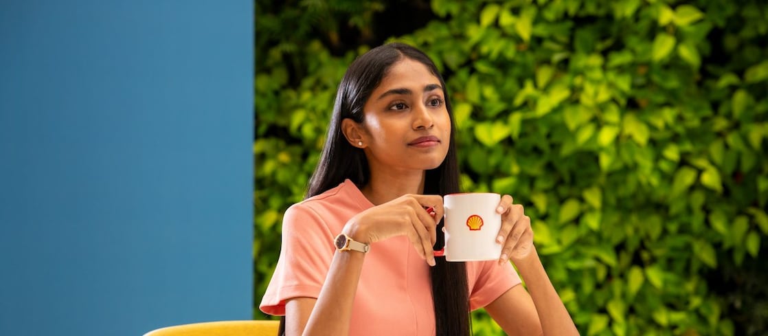 A person is holding a white coffee mug with the Shell logo, seated at a round table. The background features lush greenery, creating a refreshing and relaxed atmosphere.