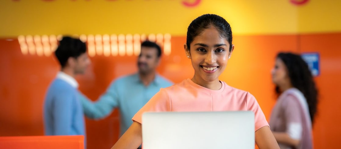 An individual is seated at a desk working on a laptop. The background shows the same orange wall with “Powering Progress” text and a group of people conversing, reinforcing the collaborative and energetic environment.