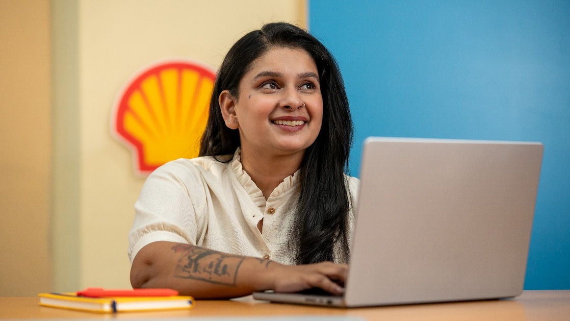 A person is working on a laptop at a desk with a Shell logo visible on the wall behind. A yellow notebook and pen are placed on the desk, suggesting a focused work environment. The background features vibrant blue and yellow panels.