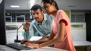 Two individuals are working together at a computer workstation. One person is seated and operating the computer, while the other is standing and pointing at the screen, suggesting guidance or explanation. The setting is a modern office with clean lines and a microphone visible on the desk, indicating a collaborative and tech-enabled environment.