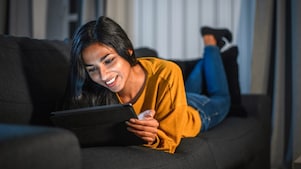 A woman wearing a yellow sweater and lying on a sofa, smiling and using a tablet.