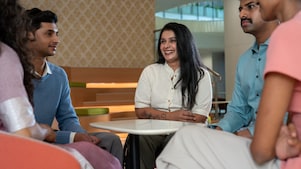 A small group is gathered around a white table in a lounge area. The setting includes patterned walls and modern furniture. One person is physically specially abled, and the group seems to be engaged in an informal discussion.