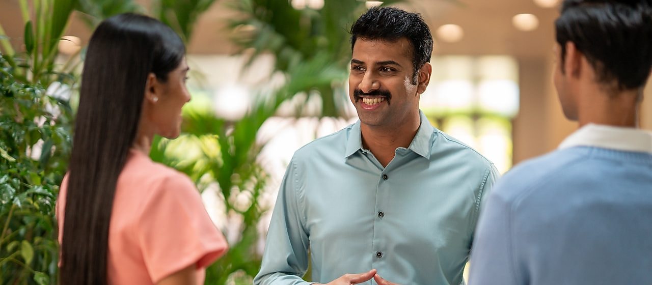 A person in a light green button-down shirt is engaged in conversation with two others. The setting appears to be an indoor space with lush green plants in the background, giving a natural and welcoming atmosphere. The individual in the center is using hand gestures, suggesting an active discussion or explanation.