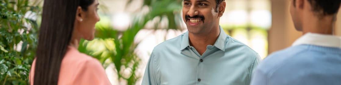 A person in a light green button-down shirt is engaged in conversation with two others. The setting appears to be an indoor space with lush green plants in the background, giving a natural and welcoming atmosphere. The individual in the center is using hand gestures, suggesting an active discussion or explanation.