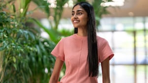 A person wearing a peach-colored outfit is standing in an indoor space with abundant greenery in the background. The setting looks bright and airy, suggesting a modern office environment with natural elements.