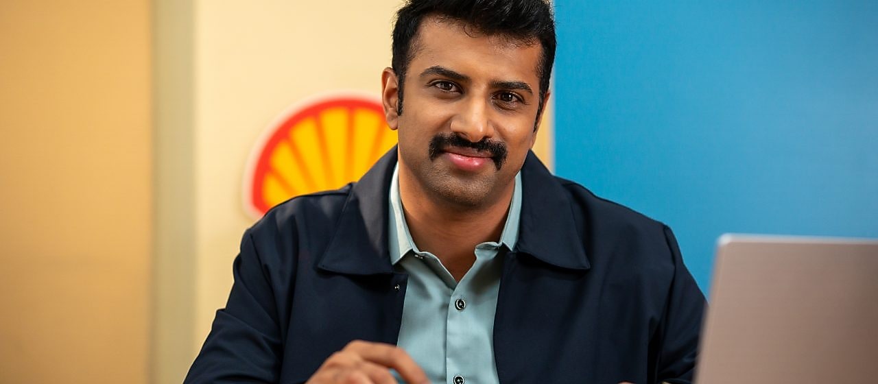 A person is seated at a desk working on a laptop, with a Shell logo visible on the wall behind. The individual is holding a pen, indicating active engagement in work or note-taking.