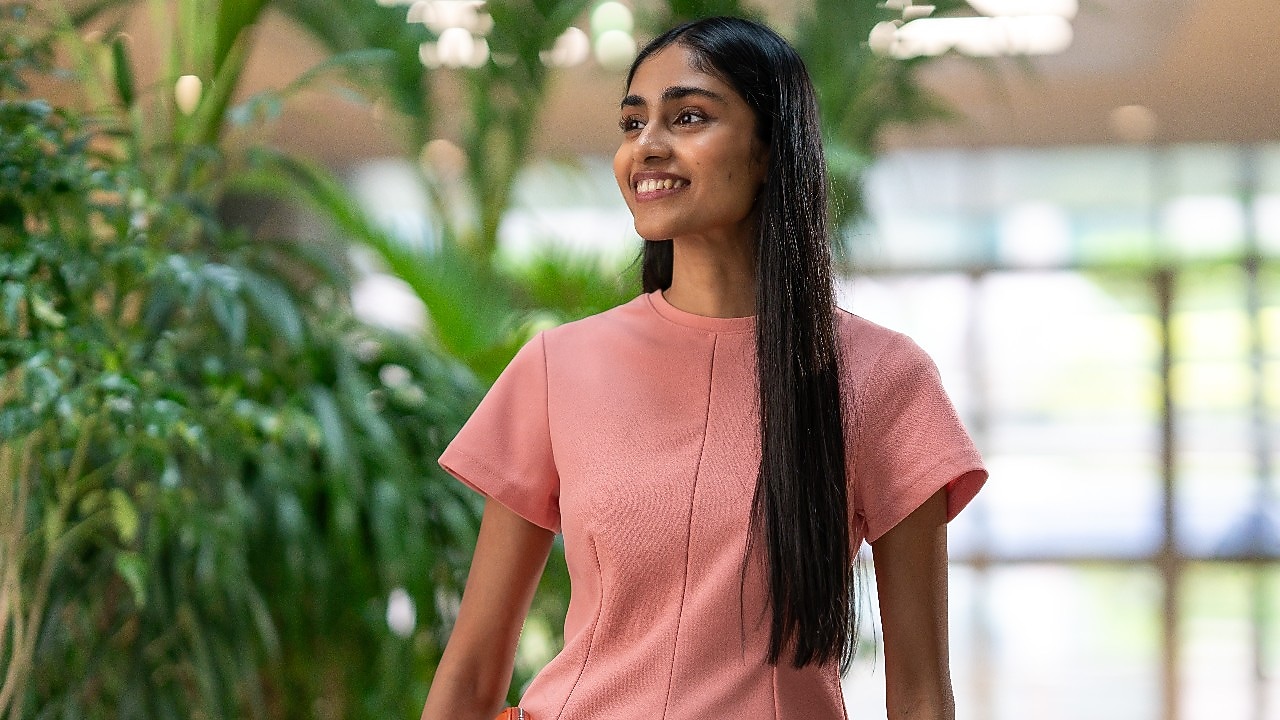 A person wearing a peach-colored outfit is standing in an indoor space with abundant greenery in the background. The setting looks bright and airy, suggesting a modern office environment with natural elements.