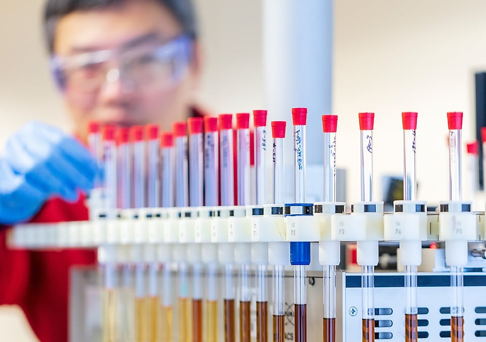 Man, in red lab coat and blue gloves handles test tubes containing fluid samples in laboratory setting.