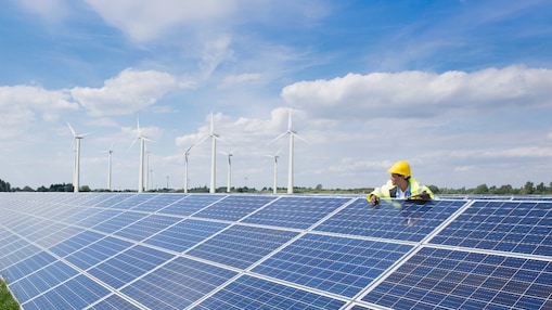 Male wearing a hardhat leaning on solar pannels with wind turbines in the background