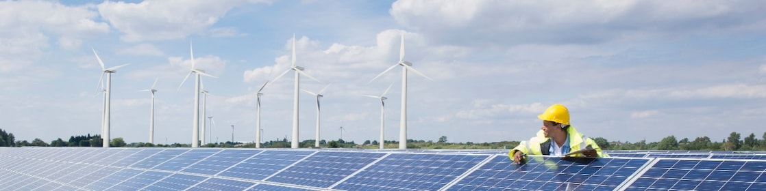 Male wearing a hardhat leaning on solar pannels with wind turbines in the background