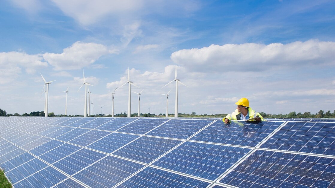 Male wearing a hardhat leaning on solar pannels with wind turbines in the background