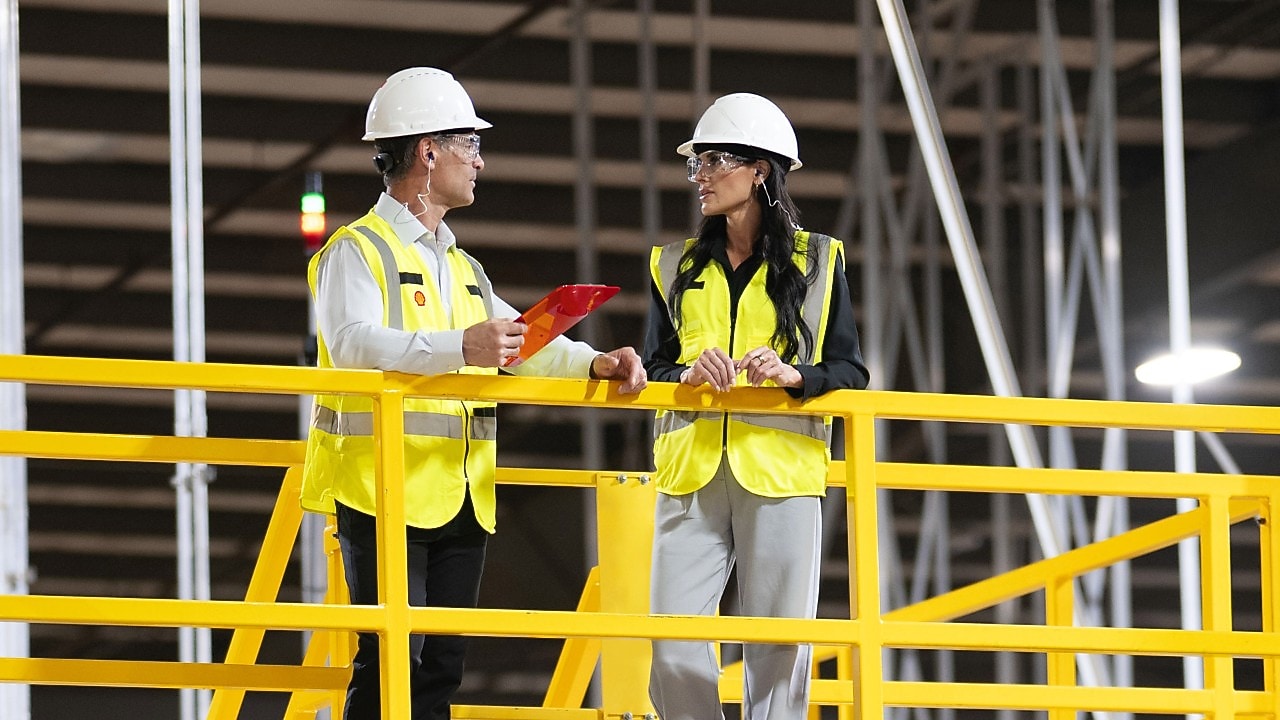 Two Shell emplyees wearing hardhats and yellow reflective jacket interacting in warehouse environment.