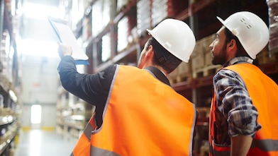 Two males wearing hardhats and orange reflective jackets in a warehouse environment. 